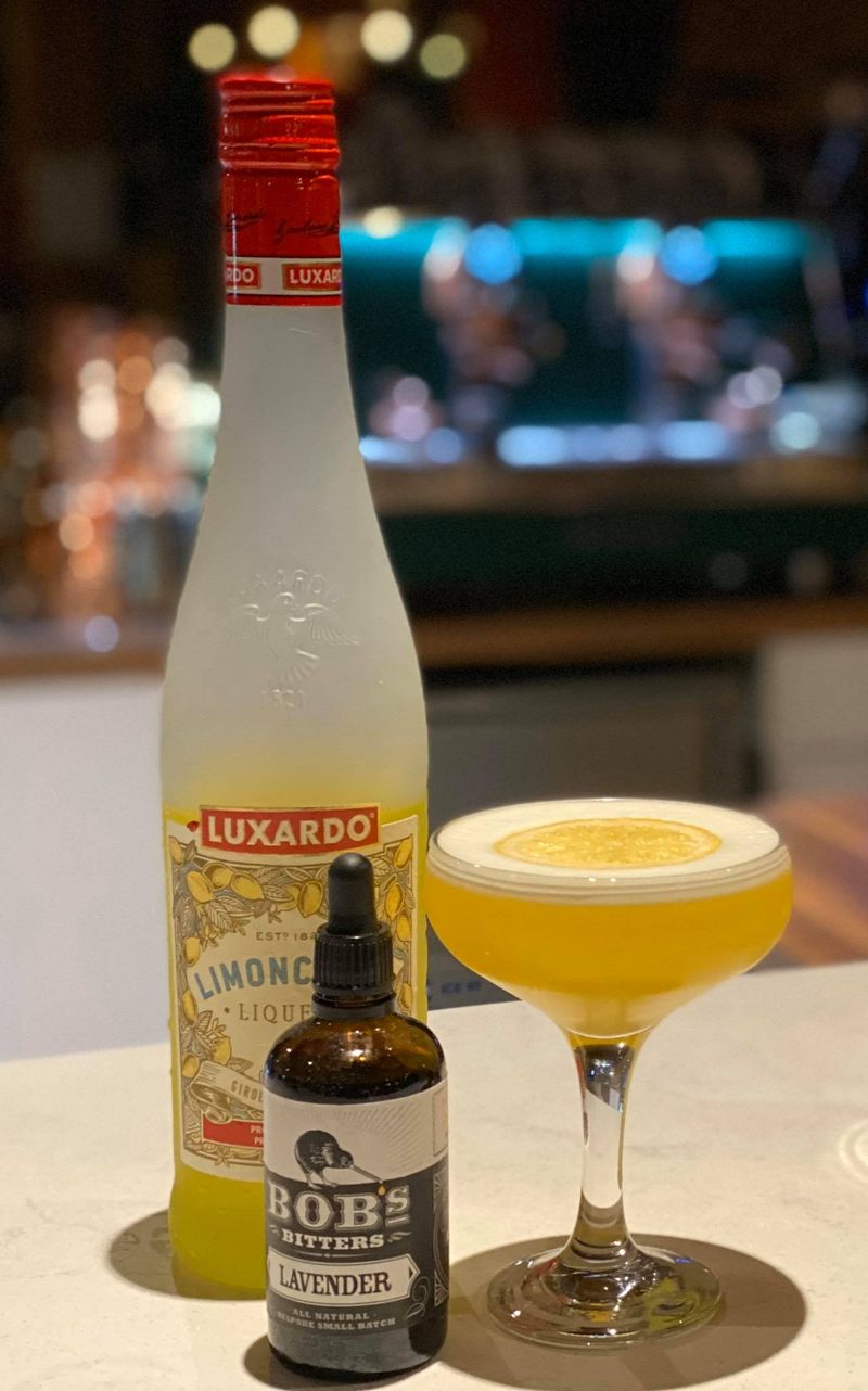 A coupe of pale yellow cocktail topped with a lemon wheel, beside bottles of limoncello and lavender bitters