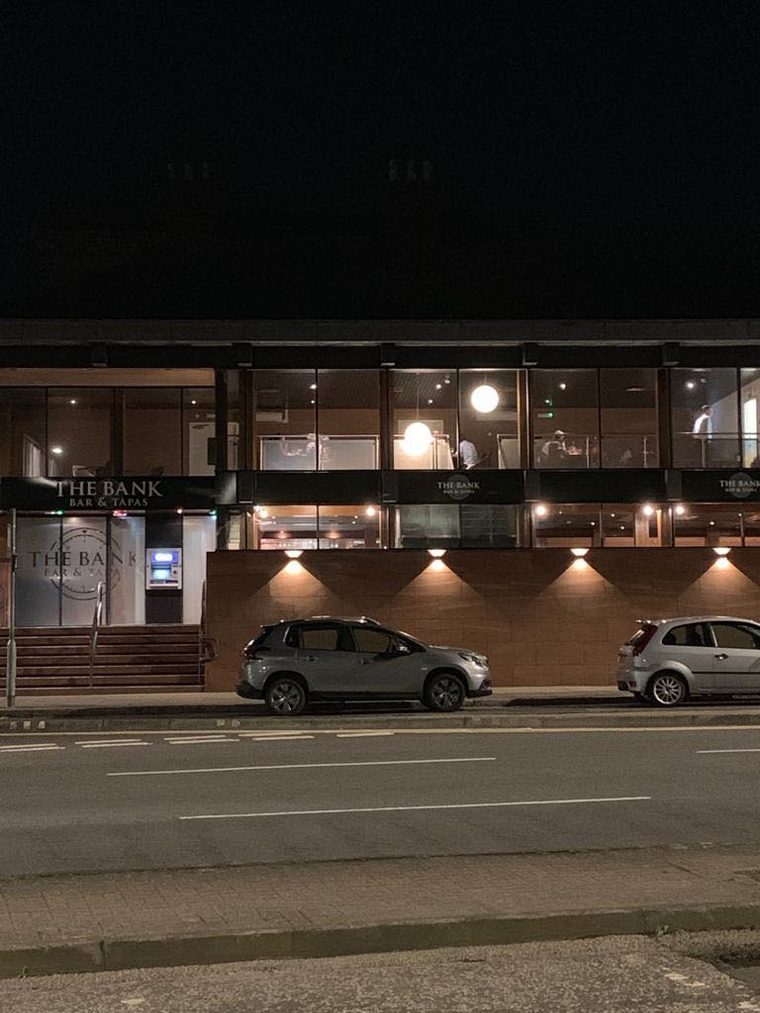 The Bank at night from across the Whitesands, its two floors of windows lit and the name in gold on the frontage