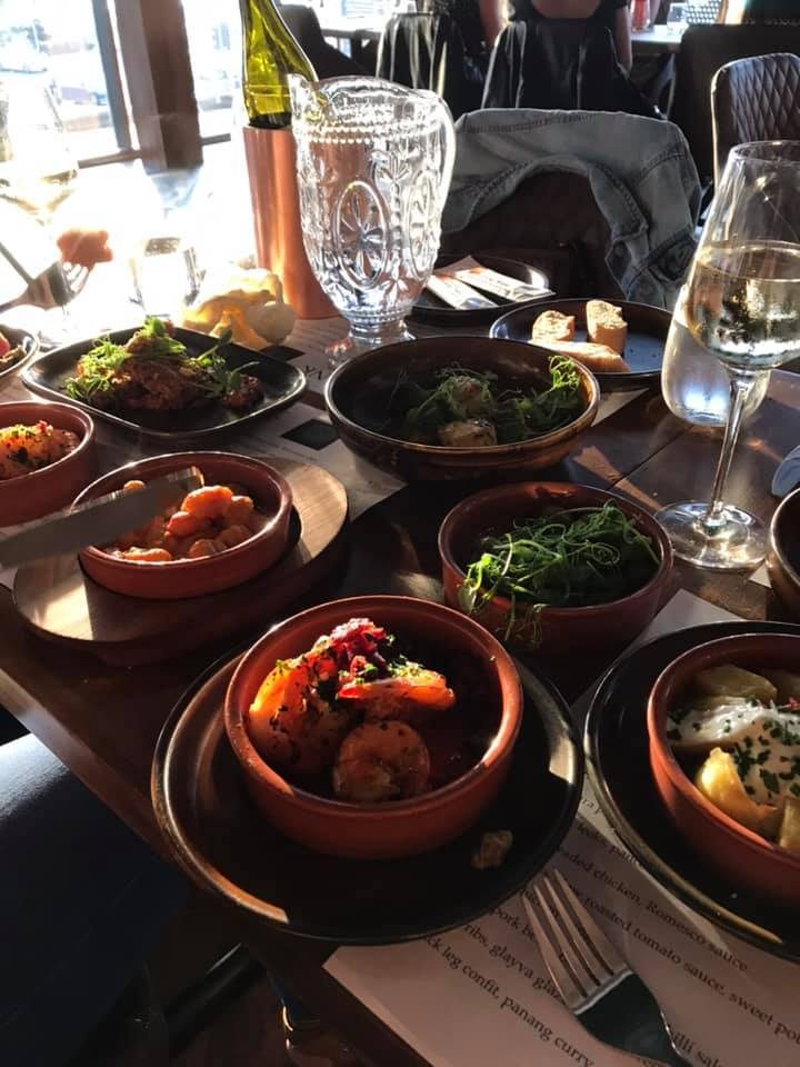 A table of tapas in terracotta dishes — prawns with chilli, chickpeas, pea shoots and bread — with white wine and a cut-glass jug